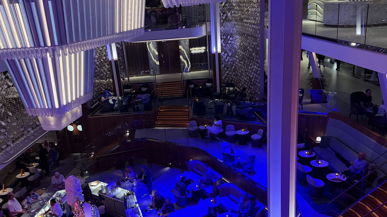 The Grand Plaza atrium at night aboard a Celebrity Edge Class ship, the LED Chandelier rising three decks above the Martini Bar