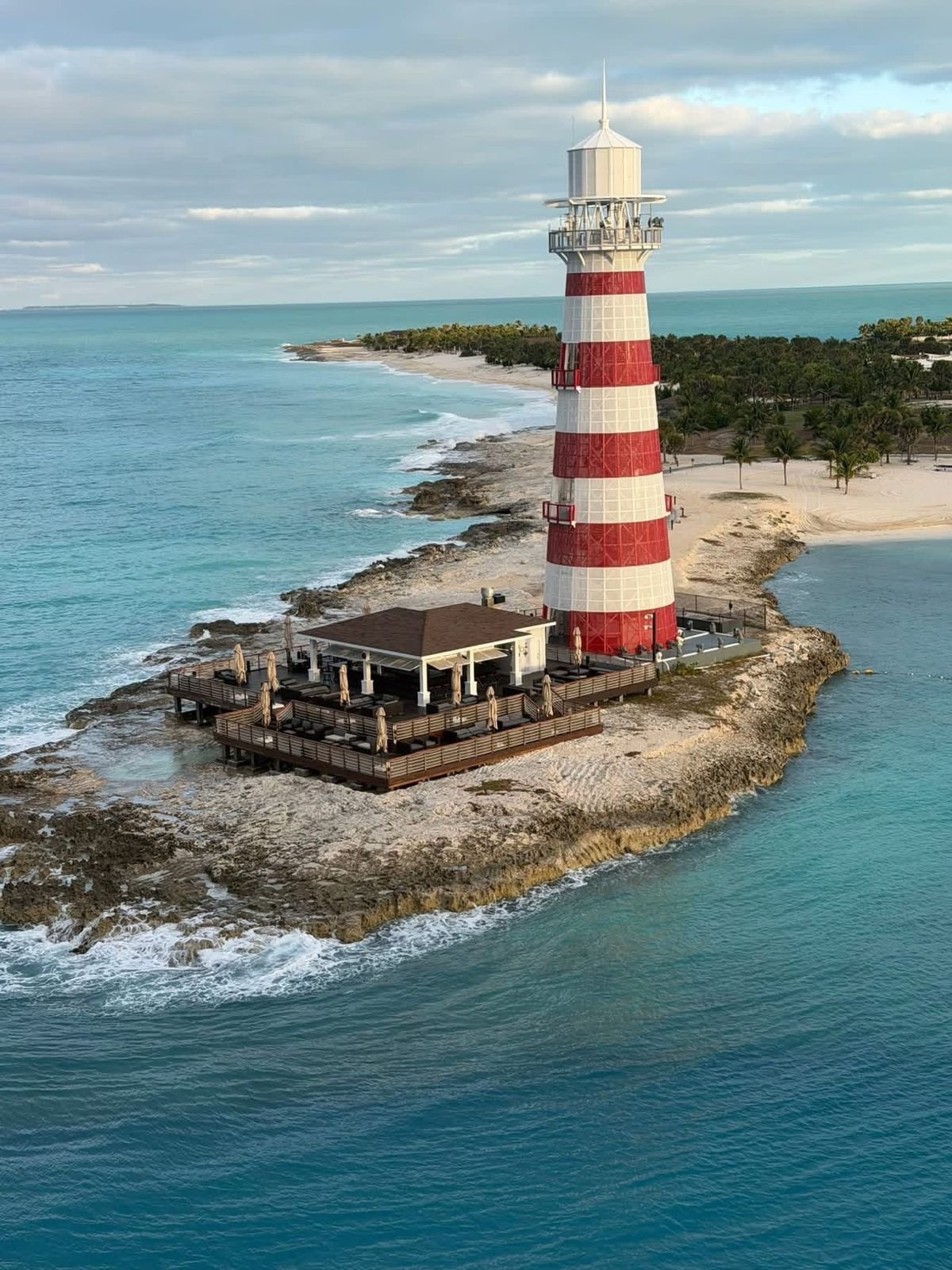 A red-and-white lighthouse on a rocky point above turquoise water, photographed from a ship
