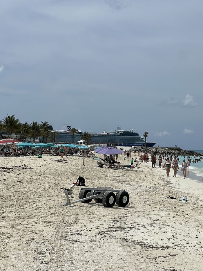 A busy Bimini beach with turquoise and purple umbrellas, swimmers in the shallows and Celebrity Silhouette docked beyond a rock jetty.