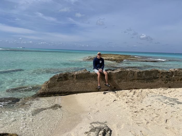 Joey Boleslawski sitting on a low rock ledge on a Bimini beach, with clear turquoise water and a reef line behind him.