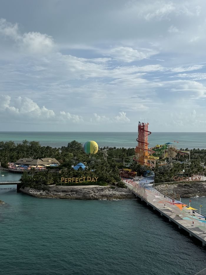 CocoCay seen from a cruise ship's upper deck: a long pier leading ashore past a large Perfect Day sign, a green and yellow balloon, and a tall red waterslide tower.