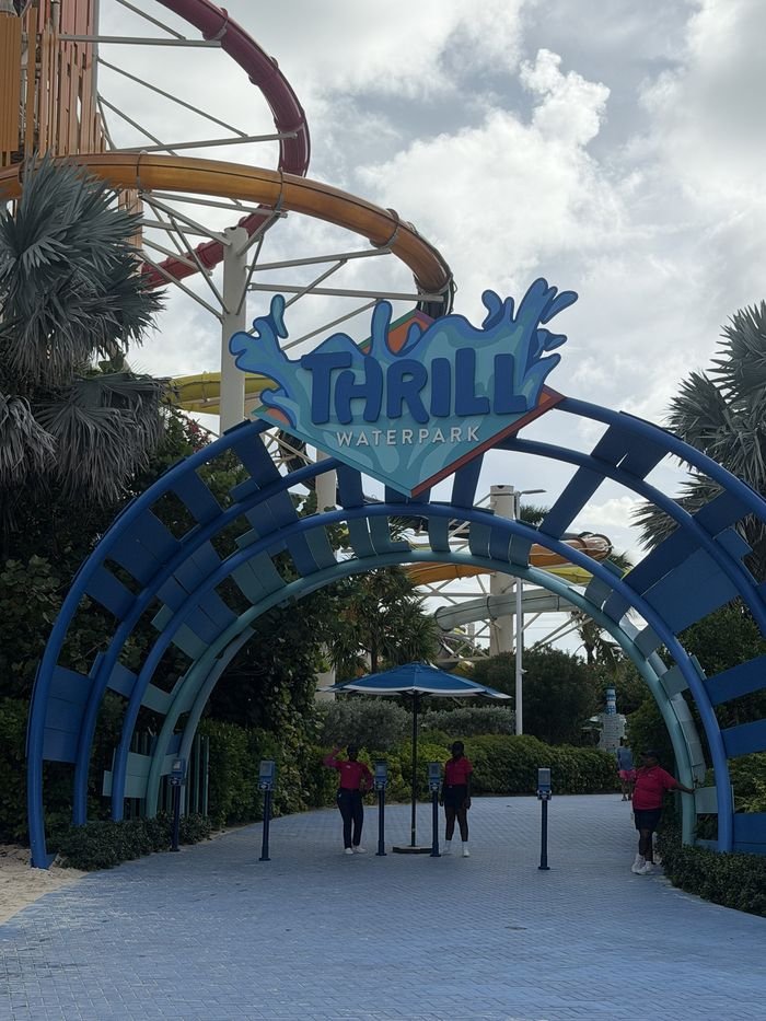 A blue arched entrance with a Thrill Waterpark sign, waterslides curving overhead and staff standing by the ticket scanners.