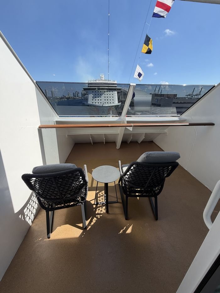A forward-facing cruise ship balcony with two cushioned woven chairs and a small round table, a solid white wall to each side and a tall glass windscreen above a wooden rail.