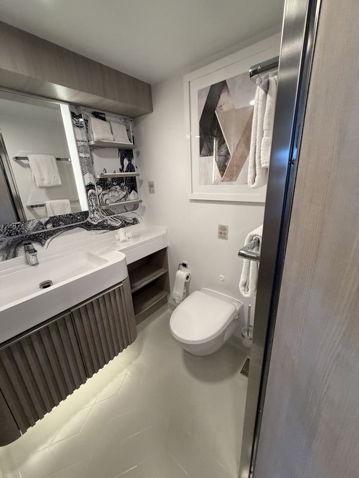 A cruise cabin bathroom with a wide white sink over a wood-slatted vanity, shelves with folded towels beside a lit mirror, and a wall-mounted toilet.