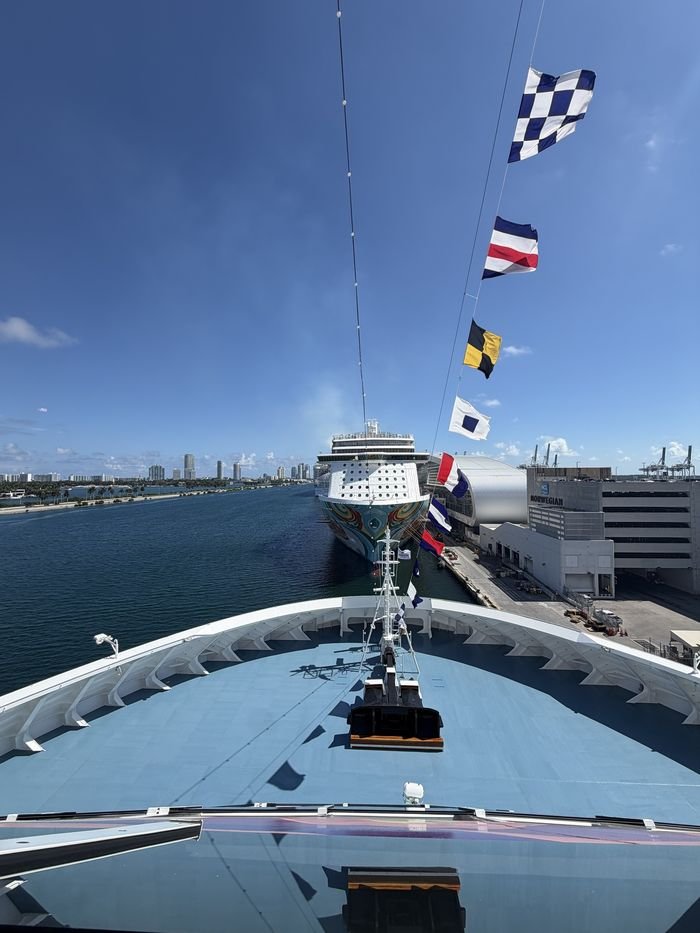 The view from a forward-facing balcony on Norwegian Aqua, looking straight over the bow at a row of signal flags, another cruise ship docked ahead and the Miami skyline across the water.