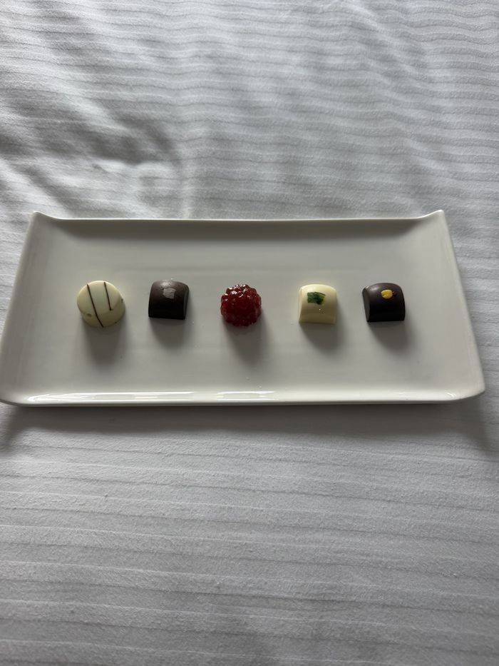 Five chocolates and a raspberry jelly lined up on a long white plate set on the bed.