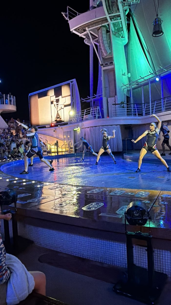 Performers in dark costumes posing on the curved stage deck of a ship's outdoor Aqua theater at night, with a large screen behind them and an audience in the stands.
