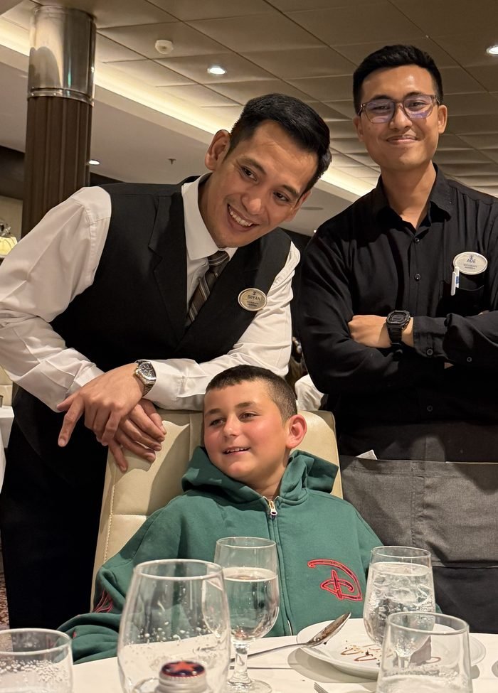 Two smiling servers in vests and black shirts with name badges stand behind a boy in a green hoodie seated at a white-clothed dining room table set with water glasses.