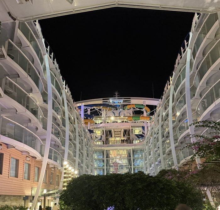A night view down the open-air center of a large cruise ship, with balconies on both sides, trees below and colorful waterslides at the far end.