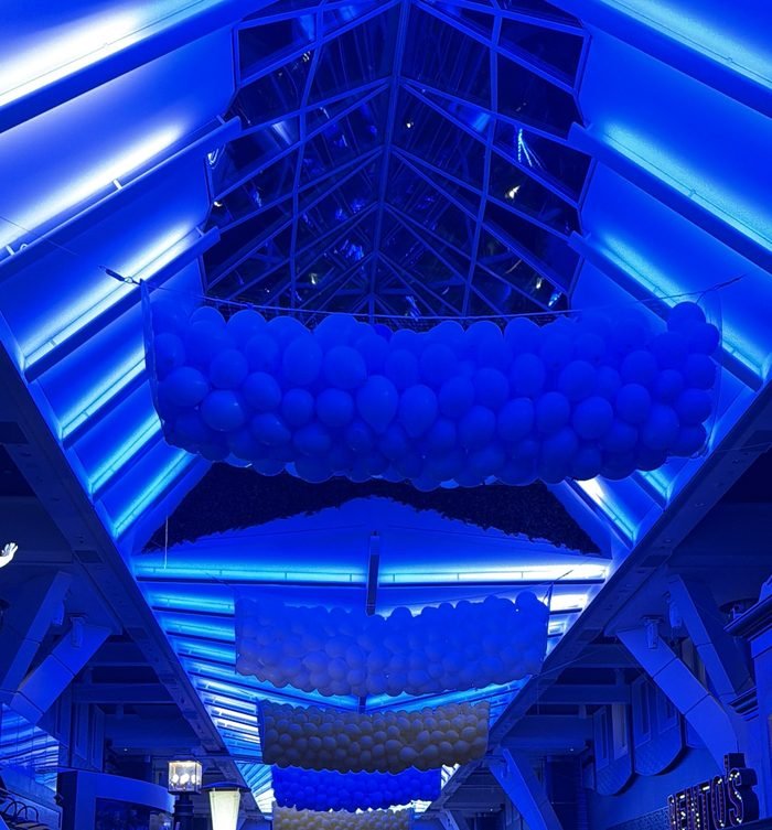 Nets of blue, white and yellow balloons hanging from the glass roof of Harmony of the Seas' Royal Promenade, lit blue.