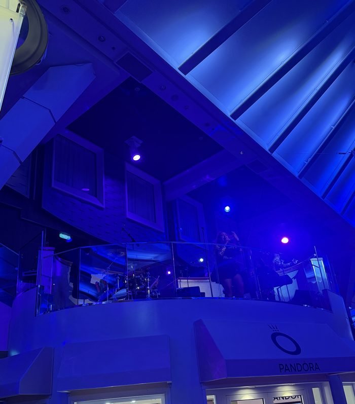 A singer and band on a glass-railed balcony above a Pandora storefront, lit blue and purple.