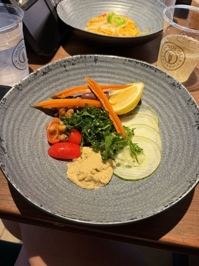 A bowl of hummus with kale, roasted chickpeas, cherry tomatoes, cucumber, carrots, olives and a lemon wedge.