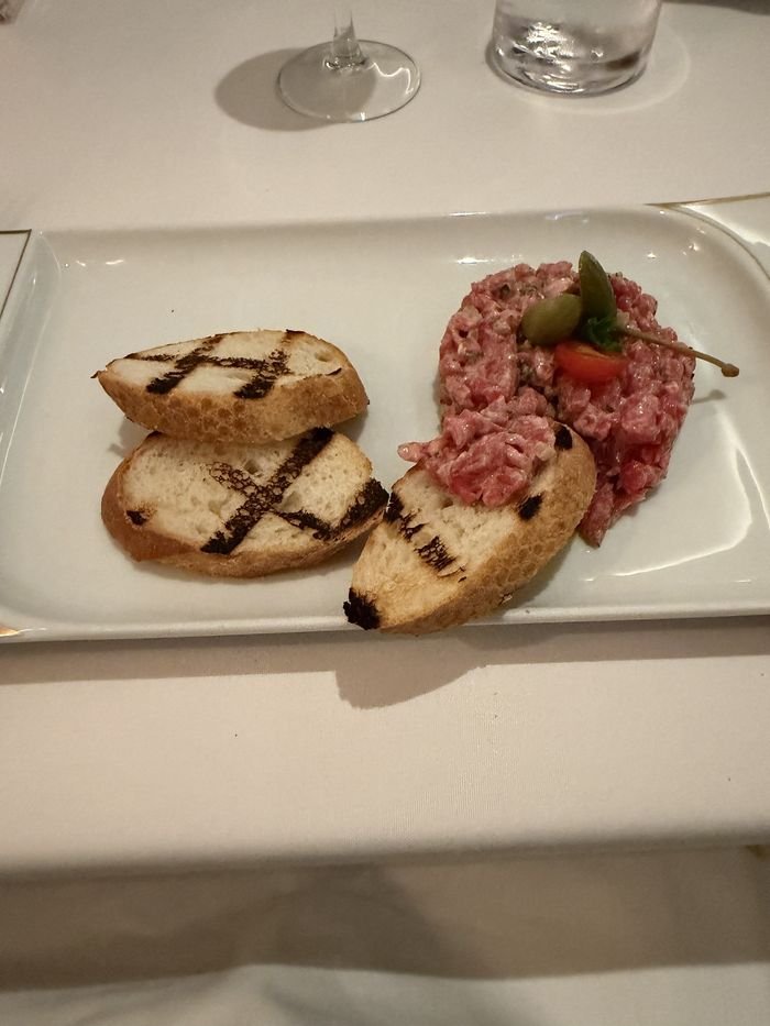 Hand-cut steak tartare topped with caper berries and a cherry tomato, beside slices of grilled bread.