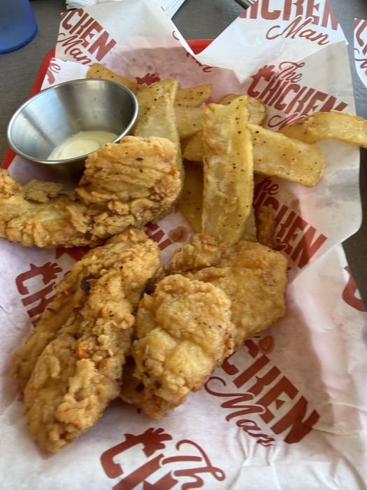 Four large golden fried chicken tenders on Chicken Man branded paper, with flat steak fries and a metal cup of white dipping sauce