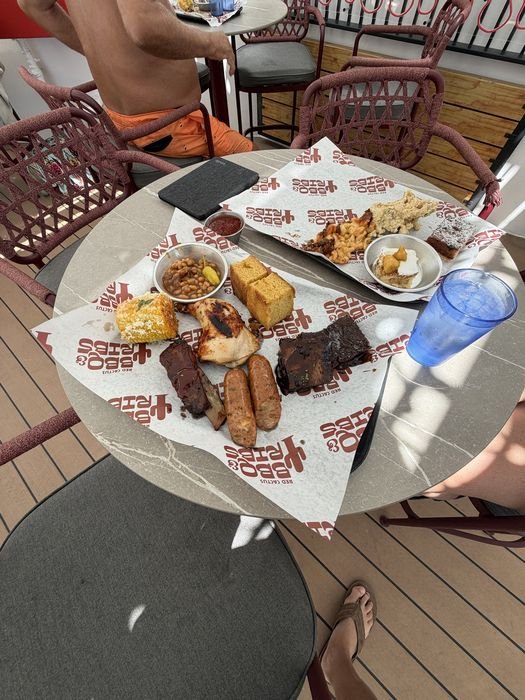 A table on the deck at Red Cactus with two trays: one of barbecue meats, cornbread, beans and corn, the other holding burnt-end mac and cheese, potato salad, banana pudding and a square of dark cake, beside a blue cup of iced water