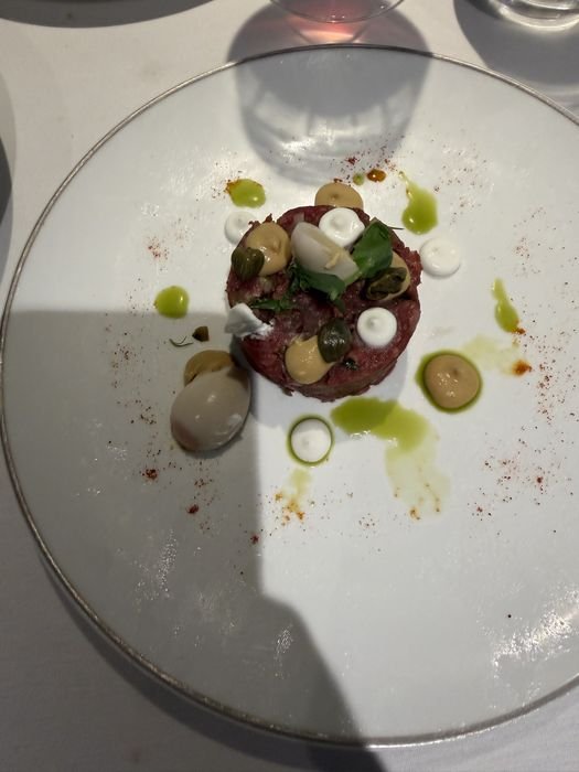 Beef tartare shaped into a neat round, topped with capers, herbs and small dots of pale sauce, with more sauce dots and green oil scattered around the rim of a white plate