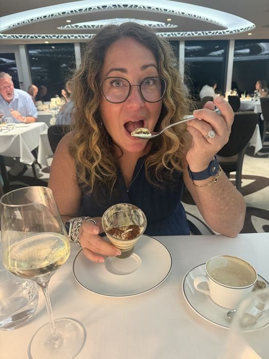 A woman in the MSC Yacht Club restaurant lifting a spoonful of tiramisu from a tall glass, with a glass of white wine and a cappuccino on the white tablecloth in front of her