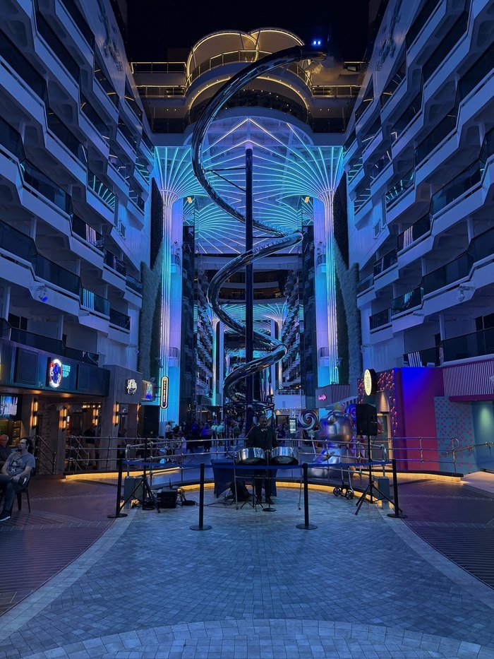 The promenade aboard MSC World America at night, lit in blue and teal, with a mirrored spiral slide winding down through the middle of the atrium and a steel pan player performing below