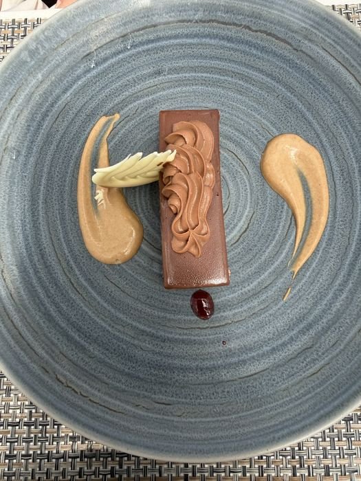 A rectangular chocolate dessert bar topped with piped chocolate mousse and white chocolate curls, plated on a blue-grey ceramic plate with two swooshes of caramel-colored sauce and a dot of red fruit sauce