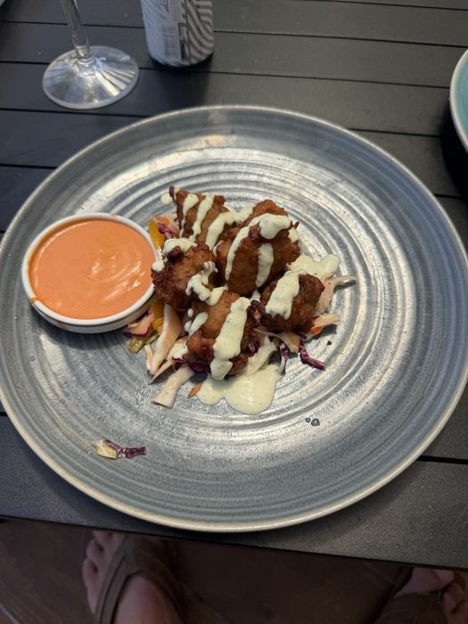 Five golden fried fritters on a bed of shredded cabbage slaw, drizzled with a pale green herb sauce, with a small dish of orange dipping sauce alongside on a grey ceramic plate