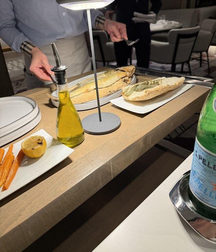 A server opening the salt crust of a whole fish on a cart, with grilled carrots, a charred lemon and a bottle of olive oil beside it.