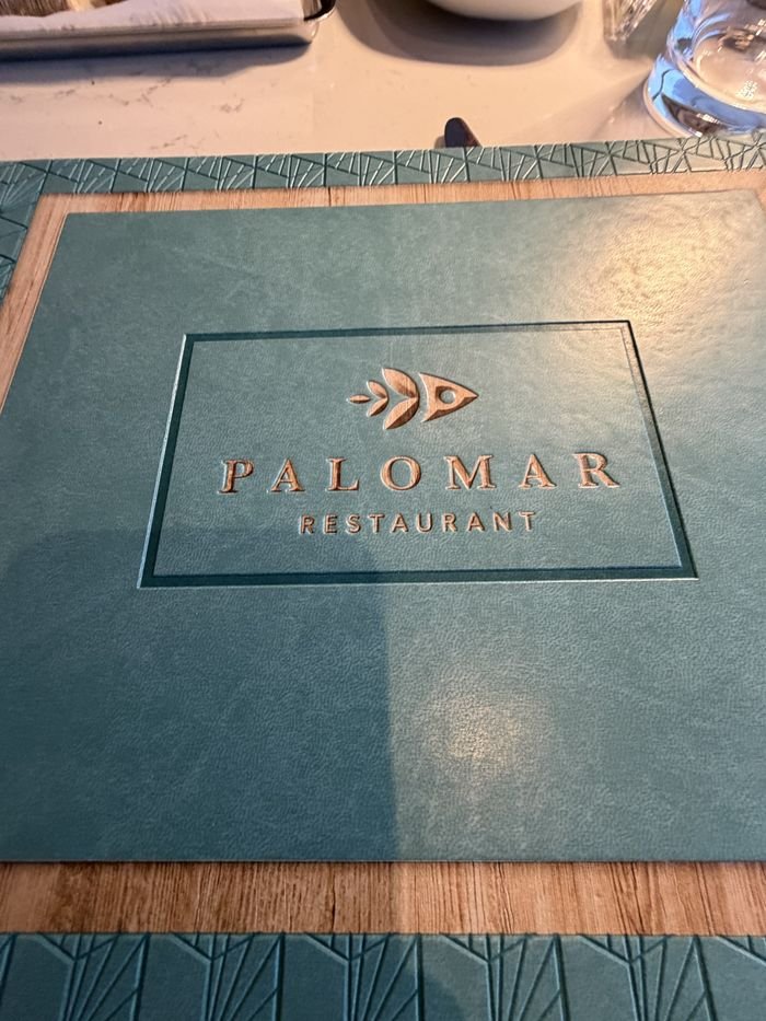 A teal Palomar Restaurant menu cover embossed with a gold fish logo, on a marble table.