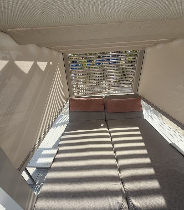 A covered two-person daybed with cream cushions and coral pillows, striped with shade from a slatted screen.