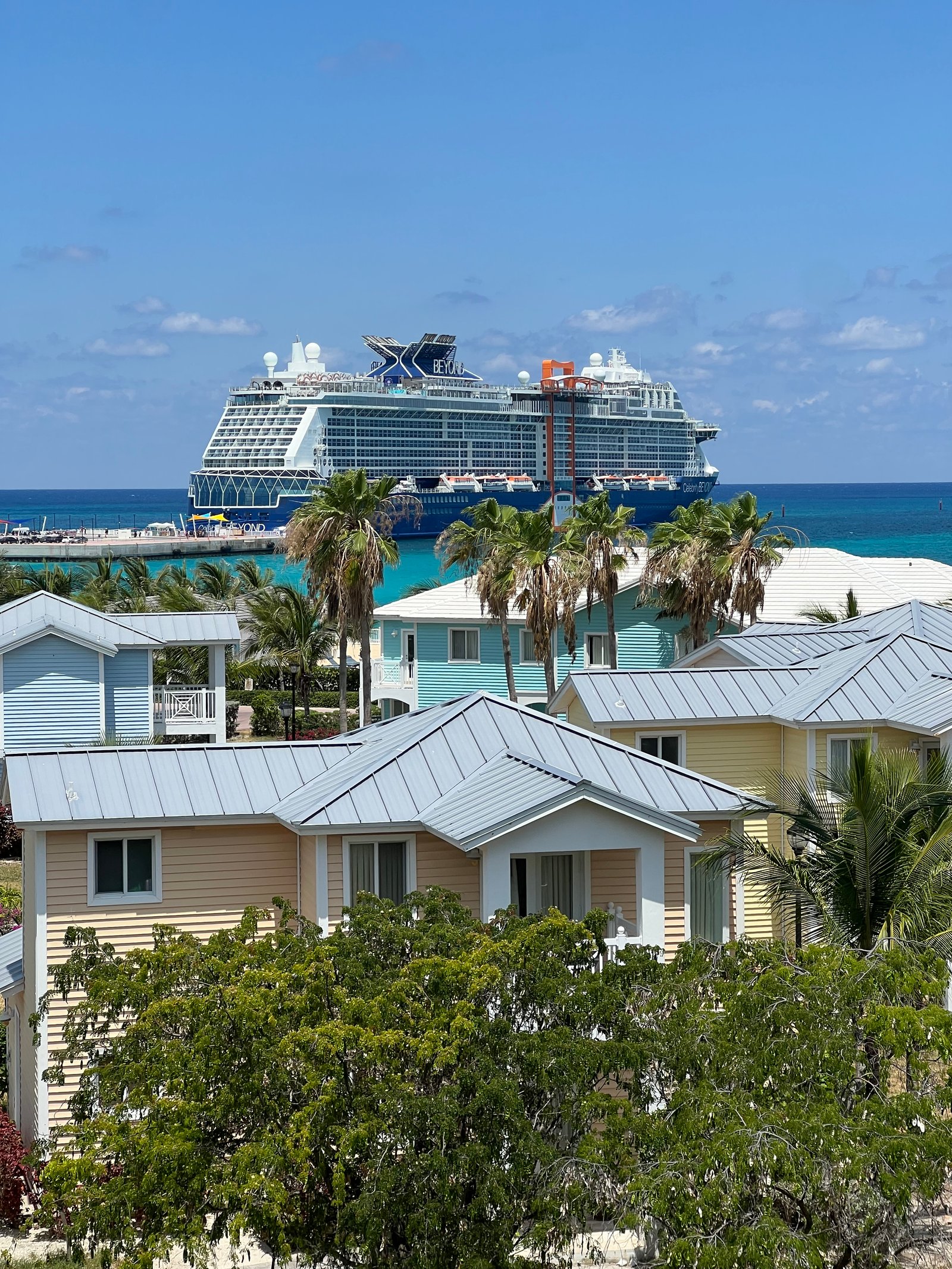 Celebrity Beyond anchored offshore at Celebrity Beach Club, Bimini, viewed past palm trees and beach cottages.