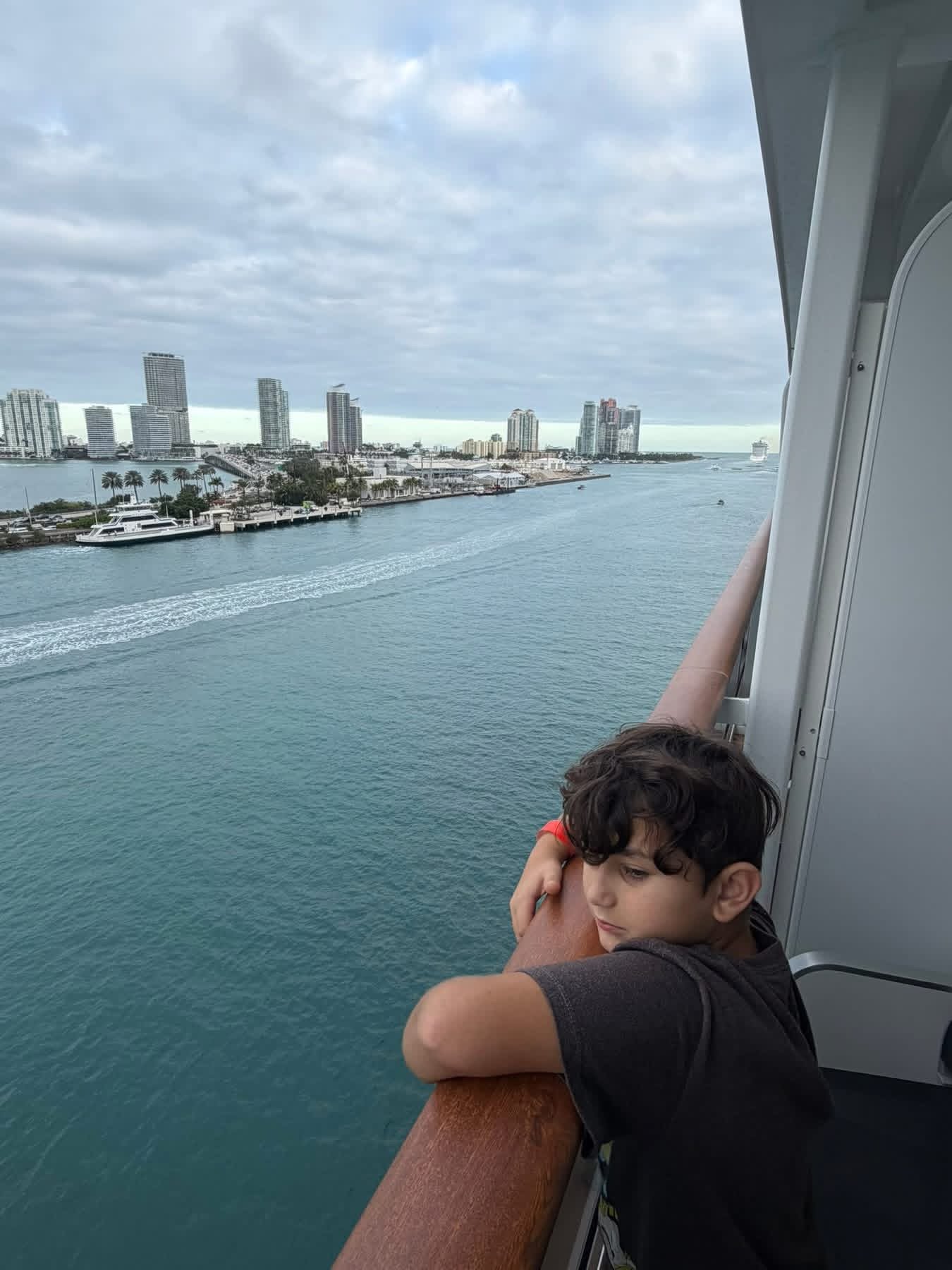 Child leaning on a cruise ship balcony railing watching the Miami skyline fade.